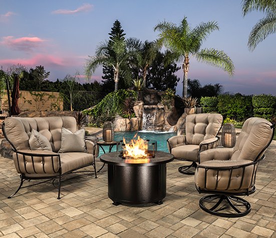 Cushioned patio loveseat and swivel chairs around a lit round fire pit beside a pool at dusk